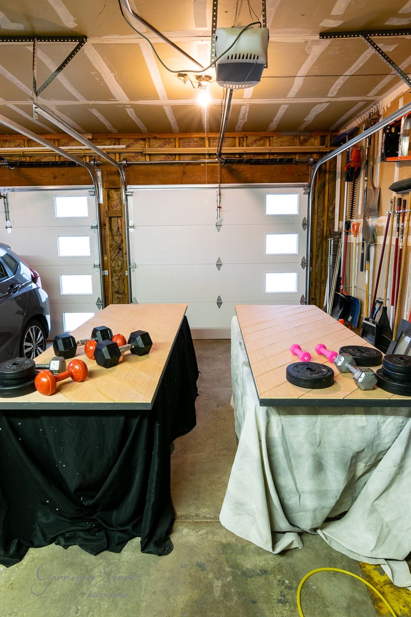 A garage converted into a gym features two workbench-style platforms. Each platform holds various weights, including dumbbells and barbells with different colored weights. A car sits to the left, while a sliding closet door adds functionality to the well-lit space under overhead lights.
