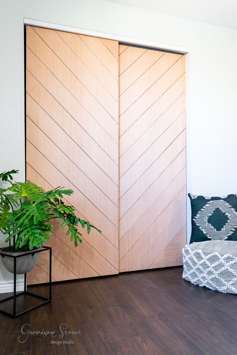 A stylish, modern interior showcases light wooden chevron-patterned sliding closet doors. A potted plant sits beside the doors, while a cushioned bench with a dark green and white patterned pillow rests on the dark wooden floor.