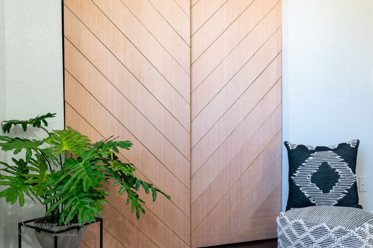 A sliding wooden herringbone-patterned closet door is on display. A lush green plant sits on a small table to the left, while a dark cushion with a white geometric design rests on a light-patterned chair to the right.