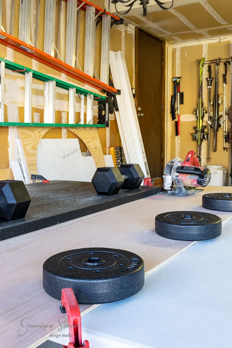 A garage workshop with a table saw and weights on a piece of wood. Shelves hold ladders, various tools, and ski poles. The sliding closet door reveals an organized, well-lit space with equipment neatly arranged for easy access.