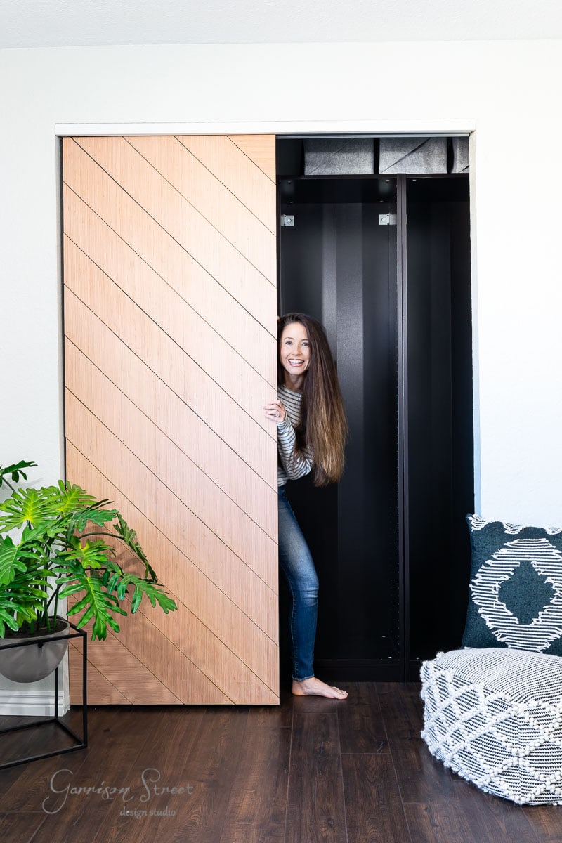 A woman peeks out from behind a partially open sliding closet door in a cozy room. The room features a green plant, patterned cushion, and dark flooring. She is smiling and wearing a striped shirt and jeans.