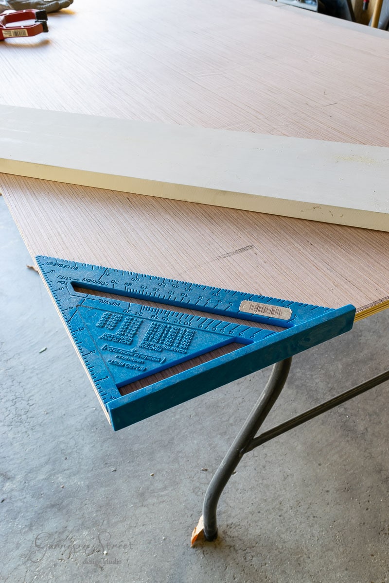 A blue speed square rests on a piece of wood placed on a table in a workshop, ready for precise measurements. In the background, a concrete floor stretches into the distance, adjacent to where sliding closet doors add practicality and sleek design to an organized space.