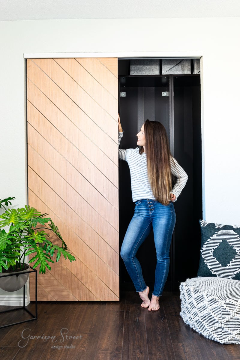 A woman in a striped shirt and jeans stands on hardwood flooring, partially opening a modern sliding closet door with a diagonal design. A potted plant and a patterned cushion add charm to the scene.