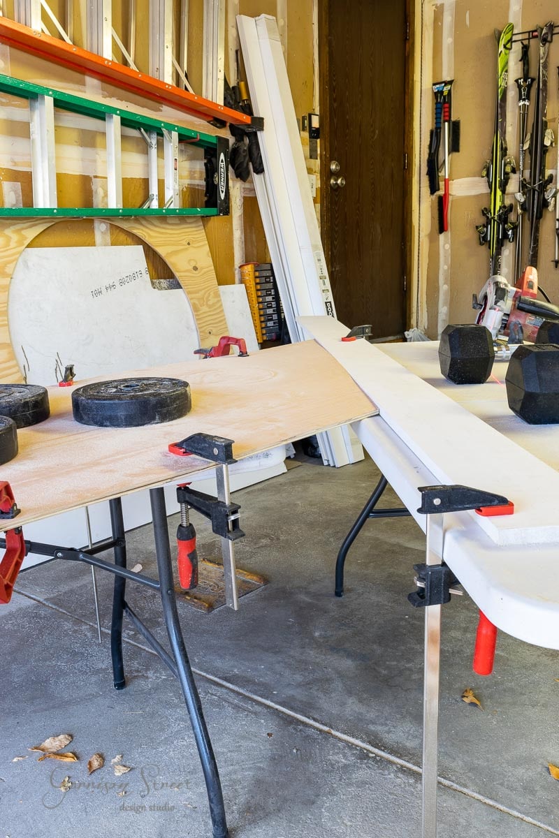 A garage workspace with a sliding sawing setup. Wooden planks are clamped onto tables, with weights and dumbbells on top. Ladders and various tools are organized along the wall, resembling a neatly arranged closet door of equipment.