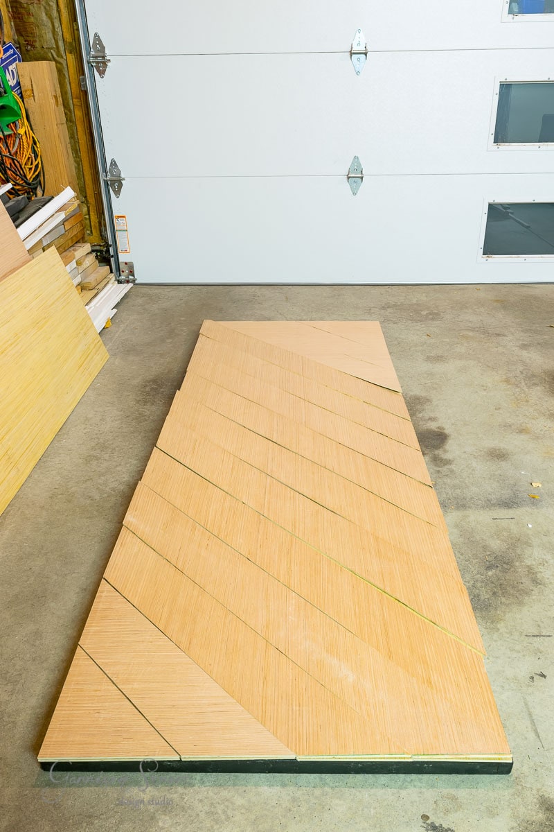 A wooden geometric pattern crafted from triangular pieces adorns the garage floor. In the background, a sliding garage door is partially open, with wooden boards and tools neatly arranged to the side.