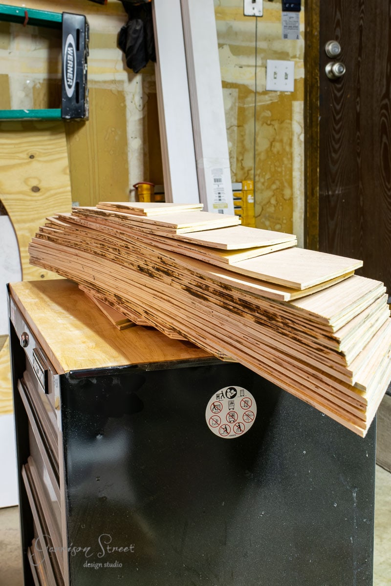 A stack of rectangular wooden boards is neatly piled on top of a black cabinet in the workshop. Various tools and materials are visible in the background, with a sliding door slightly ajar to the right.