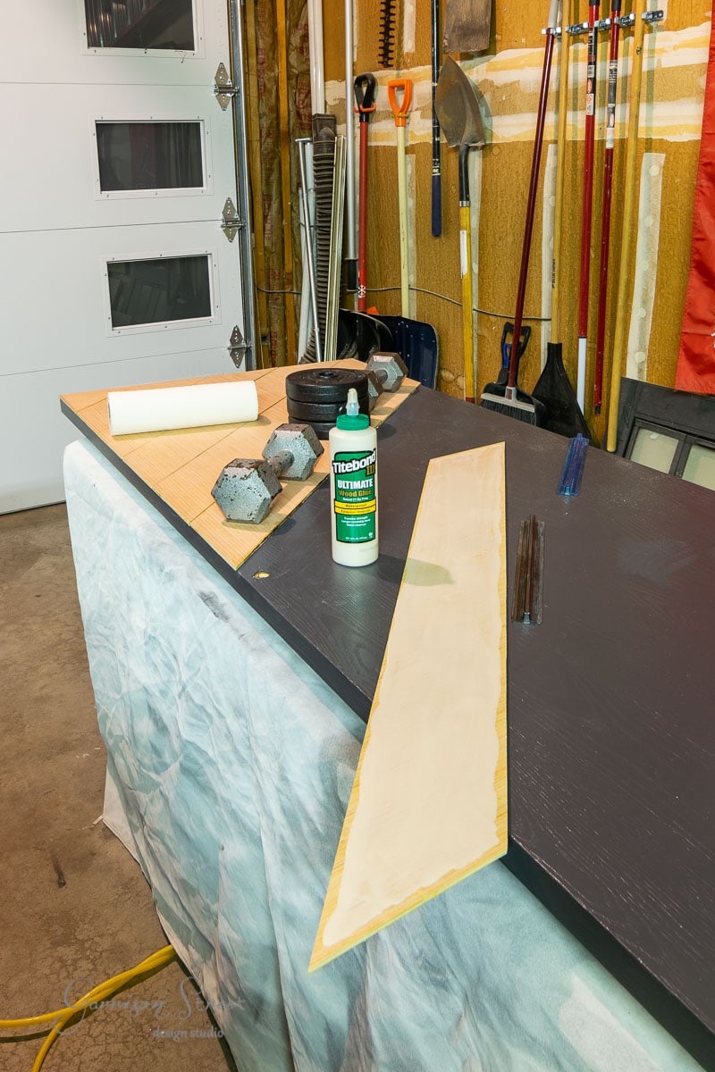 A garage workspace with various tools, including brooms, a saw, and weights. A partially painted black sliding wooden board rests on a table covered in a drop cloth. A bottle of Titebond wood glue is on the board.