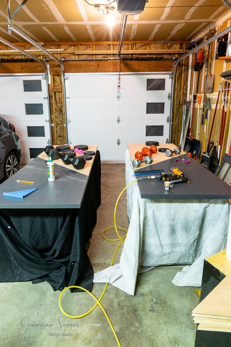 A garage workspace features two tables draped in black-and-white cloths, laden with tools, weights, and spray paint. A car is parked on the left, while sliding garage doors and walls form the backdrop.