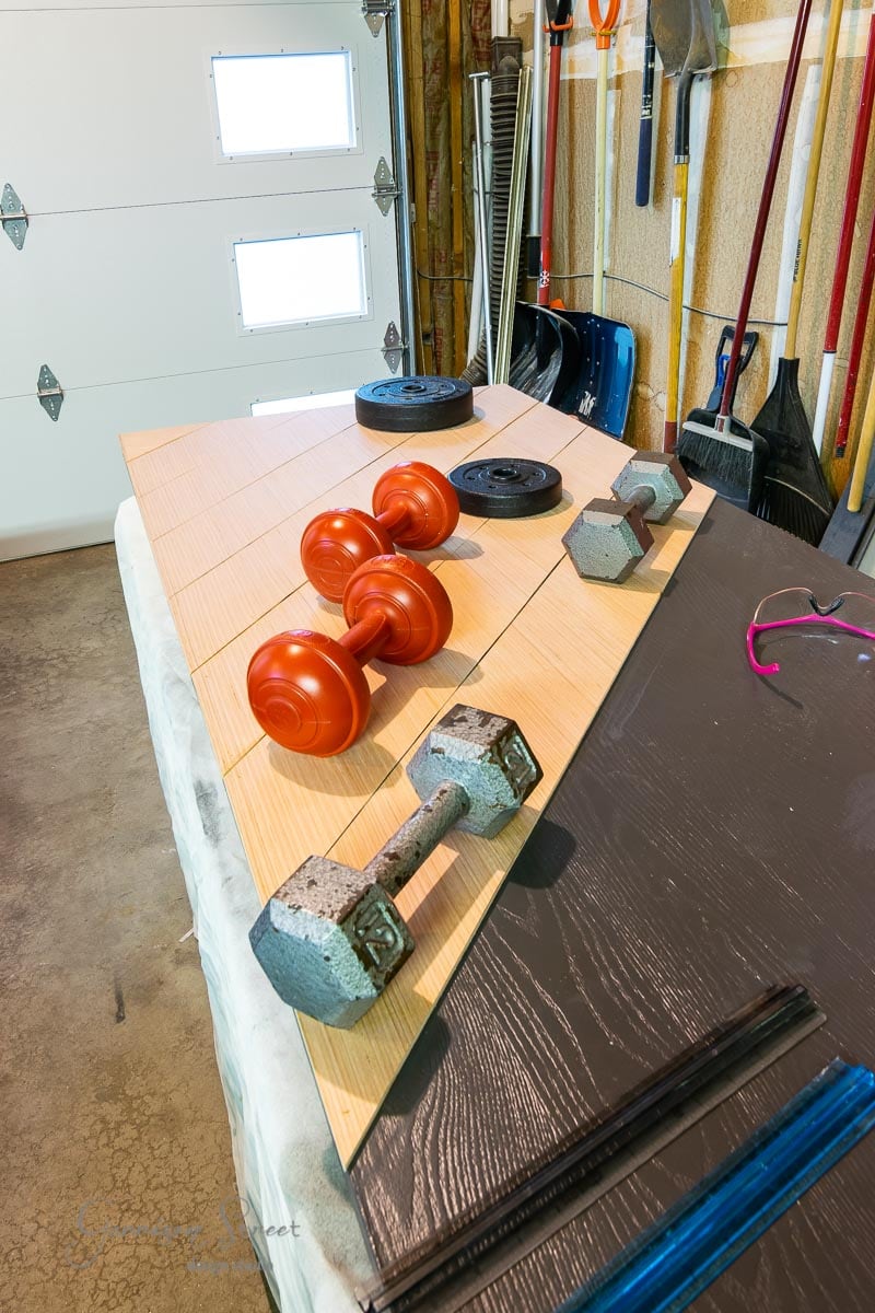 Weights and dumbbells are lined up on a piece of wood, next to a garage wall adorned with various gardening tools like shovels and rakes. The sliding garage door is partially visible in the background.