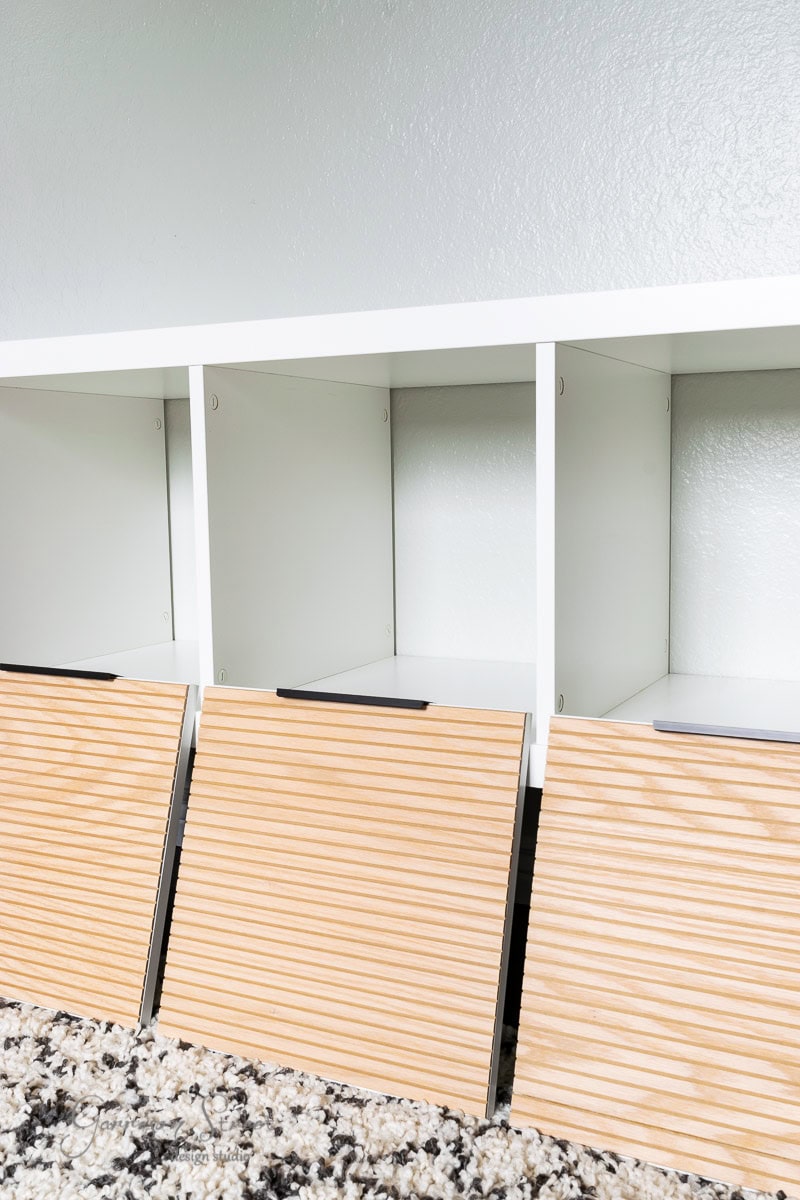 A minimalist white shelf reminiscent of an Ikea Kallax design, featuring three cubby spaces with wooden storage bins and horizontal slats. The hack includes a speckled carpet below, creating an empty yet organized look.