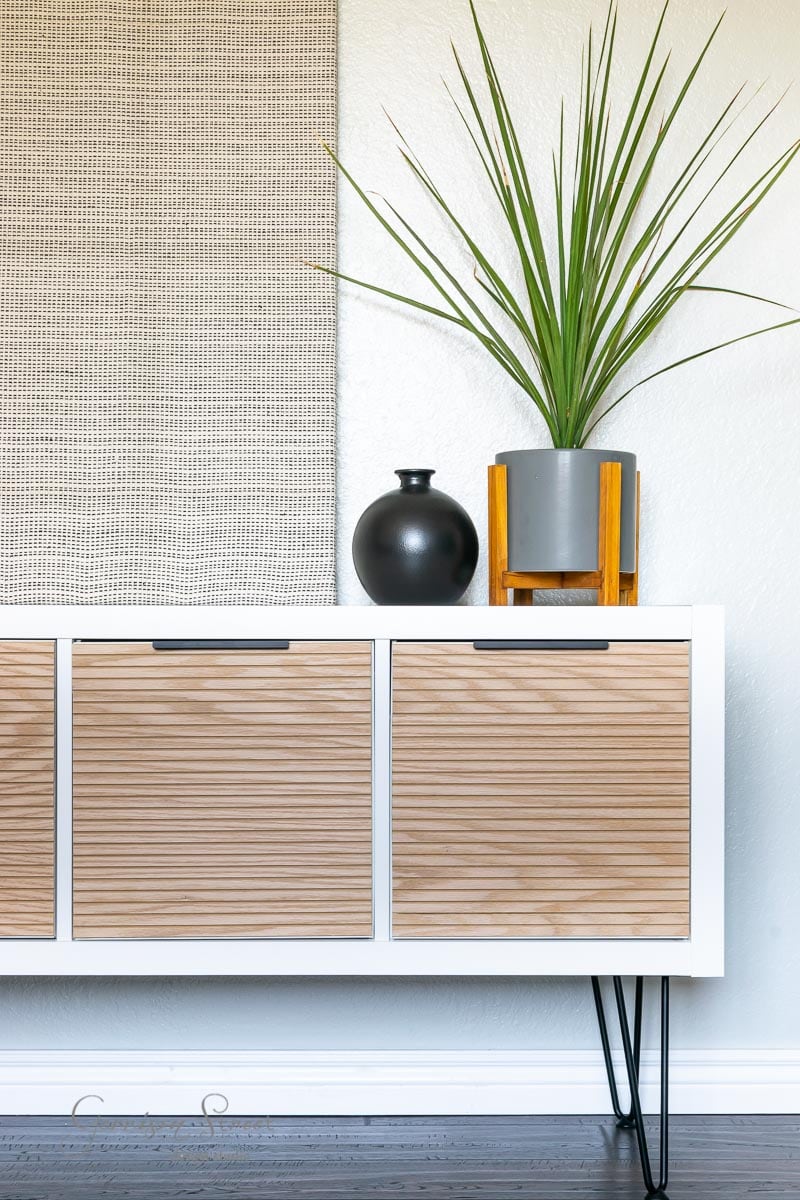 A modern room showcases an Ikea Kallax hack with white slatted doors and black hairpin legs. Atop the console, there's a black vase and a tall green plant in a gray pot with a wooden stand. A textured beige wall hanging serves as the backdrop.