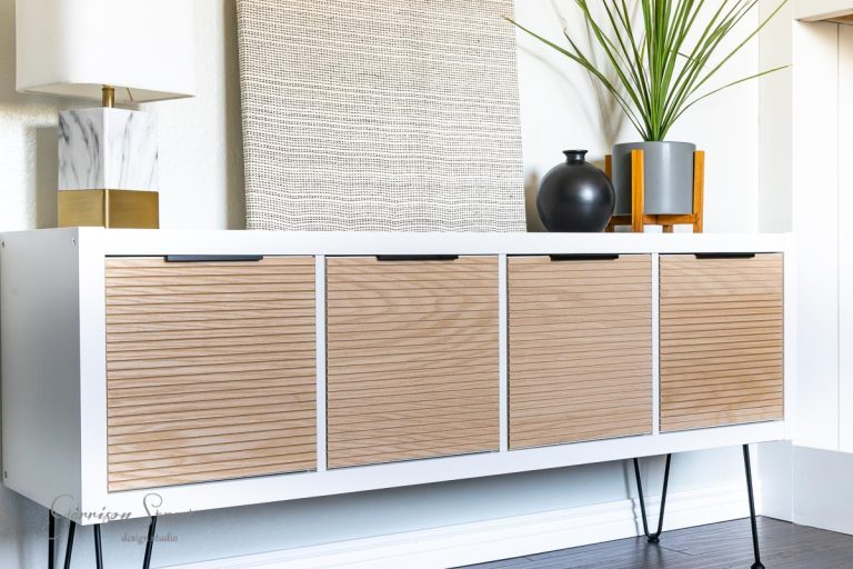A modern sideboard with ribbed wooden doors and sleek black hairpin legs sits against a wall, masterfully crafted as an IKEA Kallax hack. On top, there's a marble lamp, a black vase, and a potted plant. A textured artwork rests against the wall in the background.