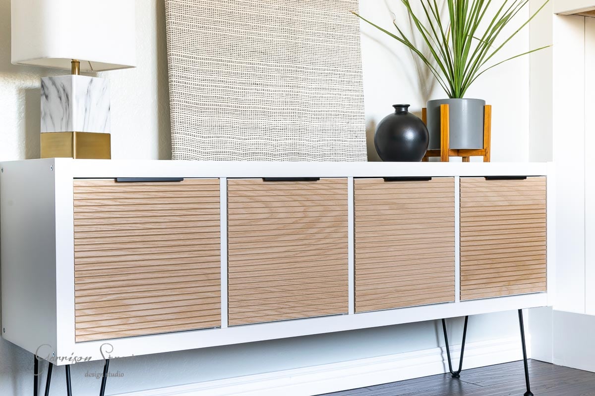 A modern sideboard with ribbed wooden doors and sleek black hairpin legs sits against a wall, masterfully crafted as an IKEA Kallax hack. On top, there's a marble lamp, a black vase, and a potted plant. A textured artwork rests against the wall in the background.