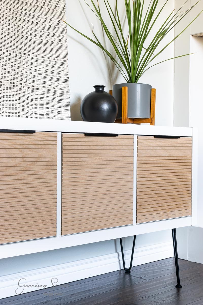 A modern sideboard with wooden slatted doors, black handles, and metal legs stands against a white wall. On top, a large green plant in a gray pot complements the round black vase. A woven wall hanging completes this stylish hack, reminiscent of an IKEA Kallax transformation.