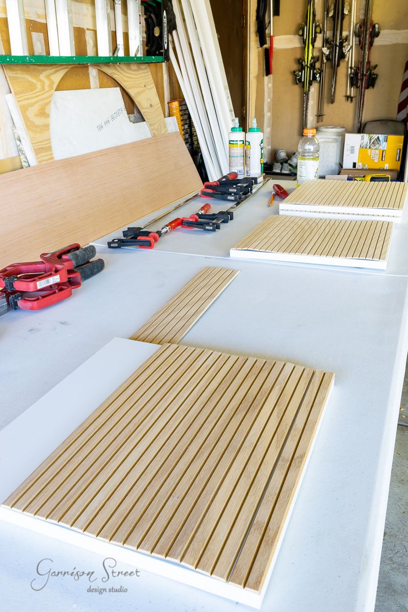 In the workshop, a table is layered with wooden slats being cleverly assembled into panels, resembling an IKEA Kallax hack. Clamps, tools, and materials are neatly arranged in this garage workspace, with various tools hanging on the wall in the background.