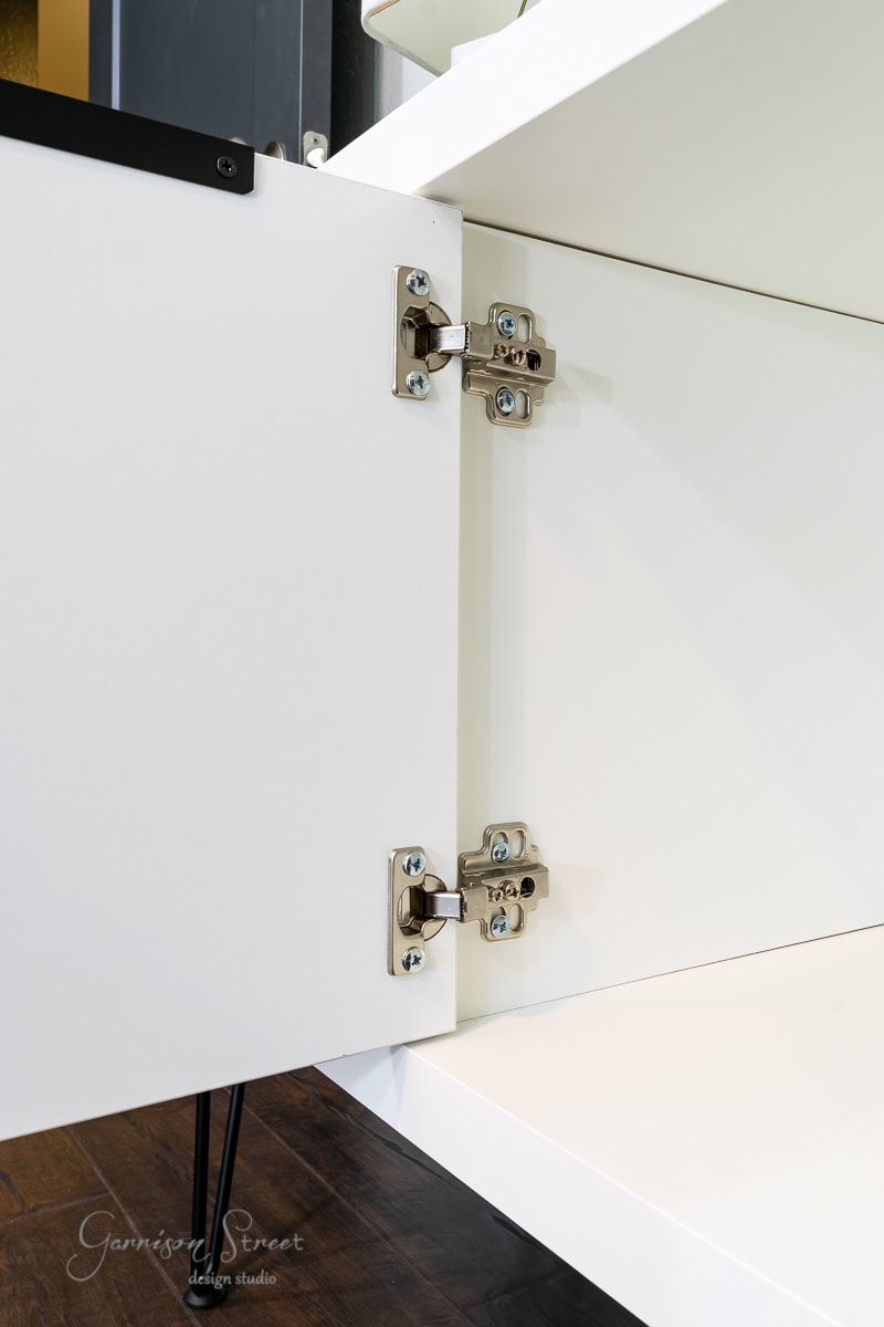 Close-up of a white cabinet door open, revealing two silver metal hinges. This stylish IKEA hack is placed on a dark wood floor, with the hinges prominently displayed, highlighting the smart design and construction reminiscent of an IKEA Kallax.