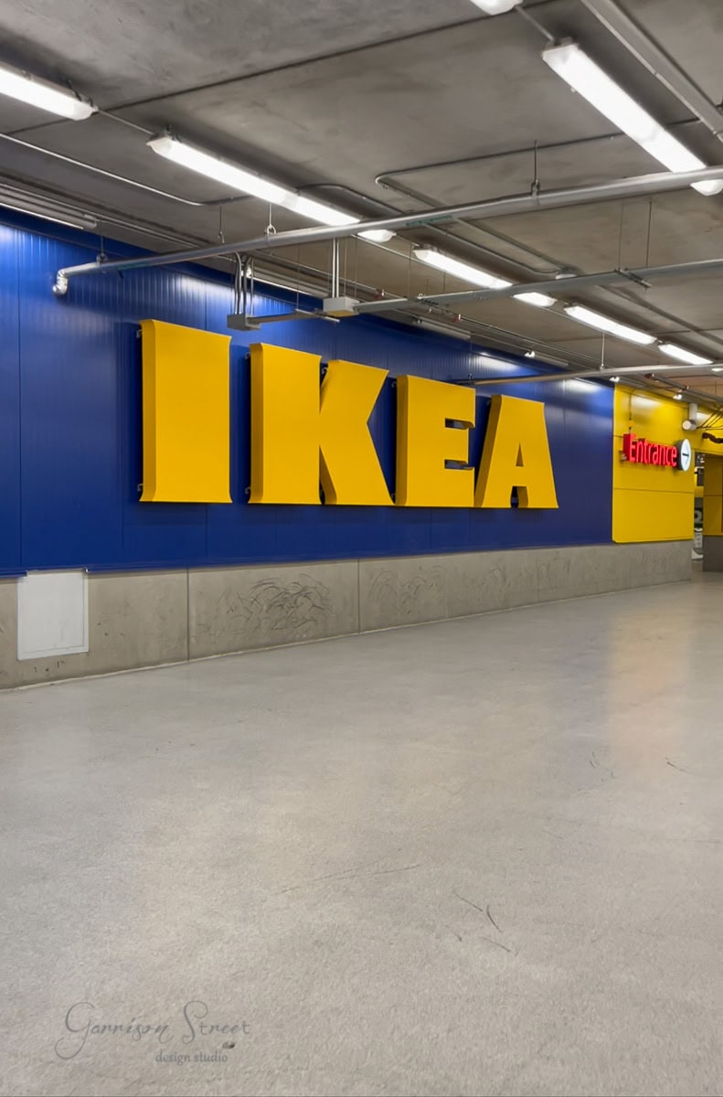 The entrance to an IKEA store features large yellow letters on a blue wall, reminiscent of the iconic IKEA Kallax design. The empty corridor with overhead lighting and a gray floor invites creative minds for a new DIY hack.