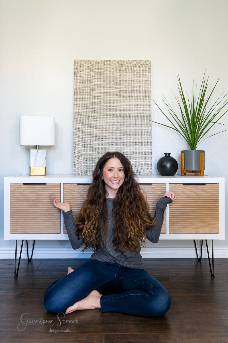 A woman with long hair sits cross-legged on a wooden floor, showcasing her IKEA Kallax hack. In front of a modern sideboard adorned with plants and decorative items, she wears a gray long-sleeve shirt and jeans, smiling with her arms slightly raised.