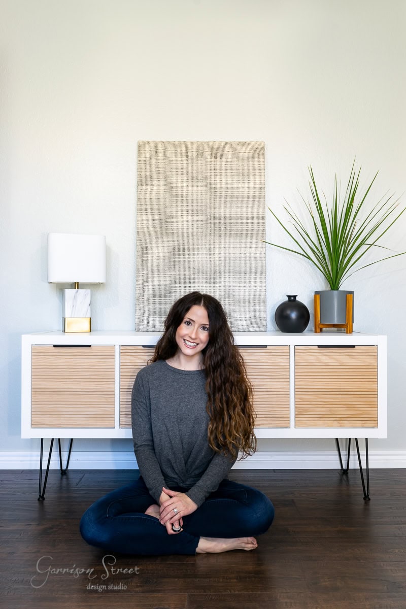 A person with long curly hair sits cross-legged on the floor, smiling. Behind them is a modern sideboard, reminiscent of an IKEA Kallax hack, adorned with textured artwork, a lamp, a small vase, and a planter with tall green leaves. The room radiates minimalist and contemporary style.