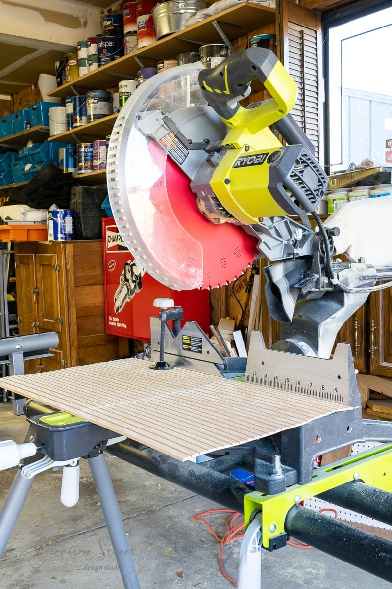 A RYOBI compound miter saw with a bright red blade is set up on a workbench in a cluttered garage, ready for your next DIY hack. Shelves filled with paint cans and tools are in the background, alongside an IKEA Kallax unit by the vented window to the right.