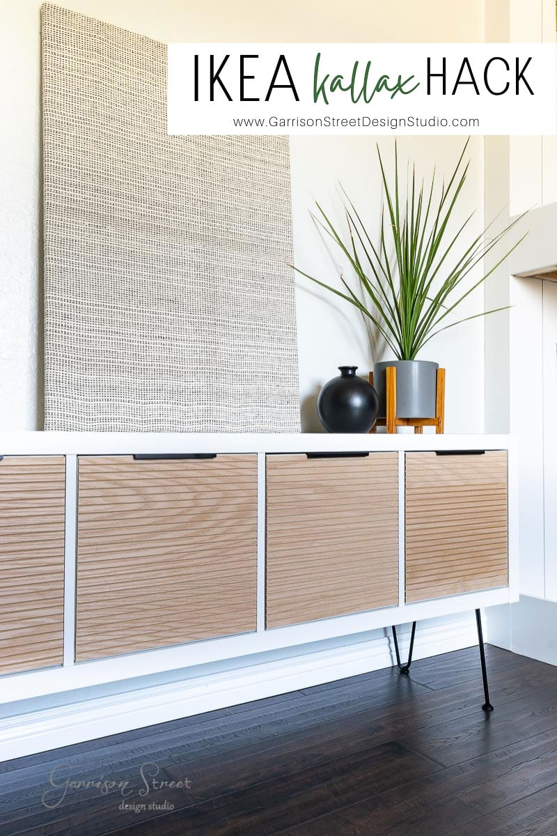 This modern sideboard is a chic IKEA Kallax hack, featuring wood-paneled doors, black legs, and a sleek white frame. Adorned with a tall plant and black vase on top, it’s perfectly complemented by a woven wall hanging in the background.