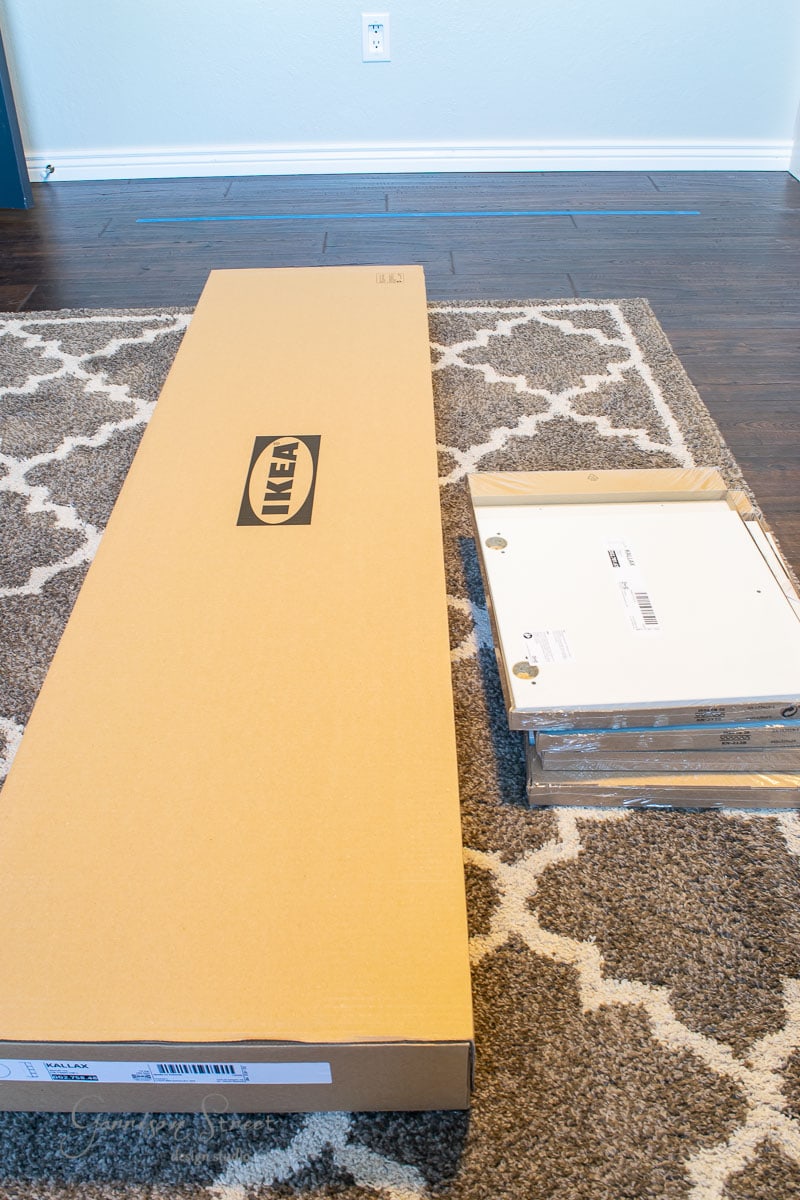 A large, flat cardboard box with the IKEA logo rests on a patterned rug next to smaller packages. Ready for an IKEA Kallax hack, they await assembly in a room with hardwood floors and a white wall in the background.