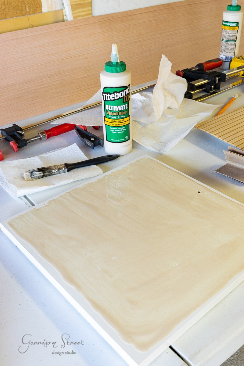 A workspace with tools, including clamps, a paintbrush, and Titebond wood glue. A piece of wood is prepared for gluing on a white surface. The scene suggests a woodworking project or an IKEA Kallax hack in progress.