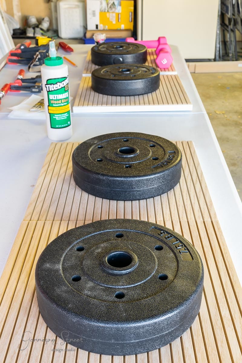 Black weight plates are arranged on wooden slats in a workshop, reminiscent of an Ikea Kallax hack. A bottle of wood glue and various tools are visible in the background, suggesting an organized setting ready for a DIY project.