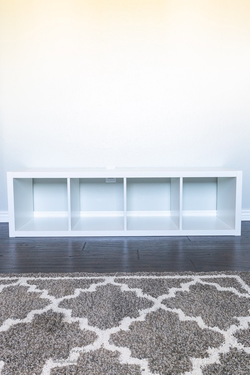 A white IKEA Kallax organizer with four cubes sits against a light-colored wall on a dark wooden floor. In the foreground, a patterned rug with a geometric design in neutral tones is partially visible—a perfect canvas for an innovative storage hack.