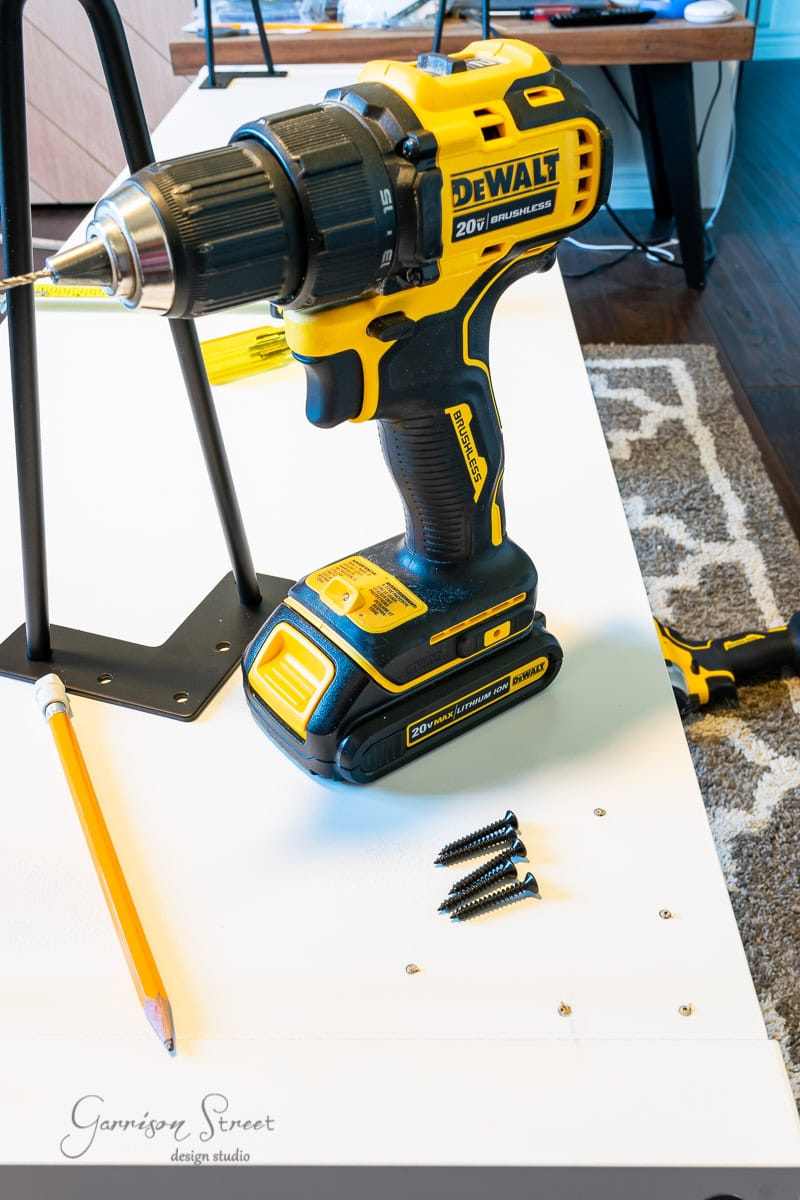 A yellow and black drill rests on a white surface next to a pencil and four screws, ready for an IKEA Kallax hack. Nearby, a partially assembled furniture piece stands with the corner of a patterned rug visible in the background.