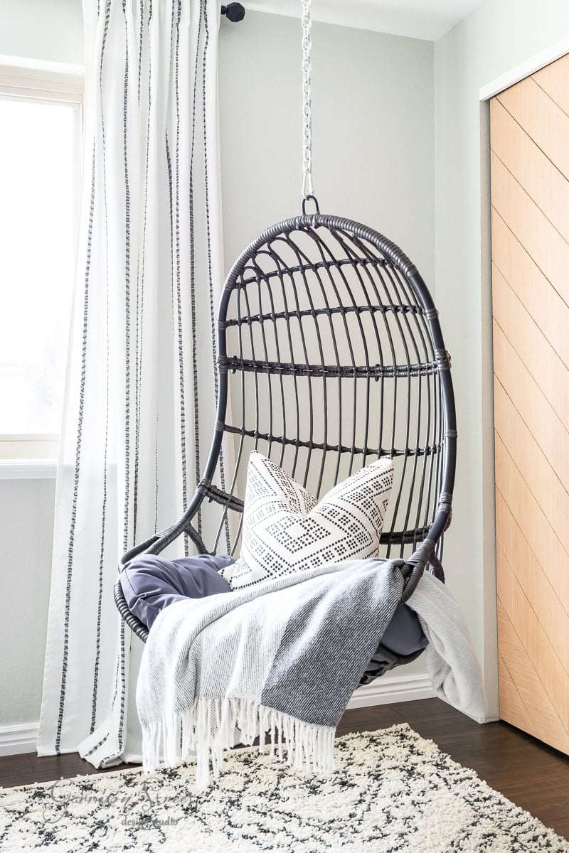 Cozy bunk room featuring a hanging rattan egg chair adorned with a geometric-patterned pillow and a soft gray throw. The space boasts striped curtains, wooden floors, and light-colored walls, crafting a serene and inviting atmosphere.