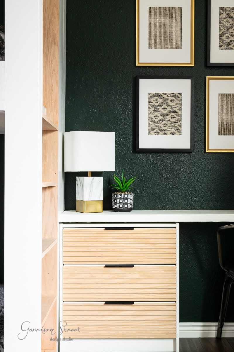 A modern home office corner nestled next to a cozy bunk room features light wood drawers, a white and gold lamp, and a small plant. Four framed geometric prints adorn the dark green wall above the desk, with a hint of shelving visible on the left side.