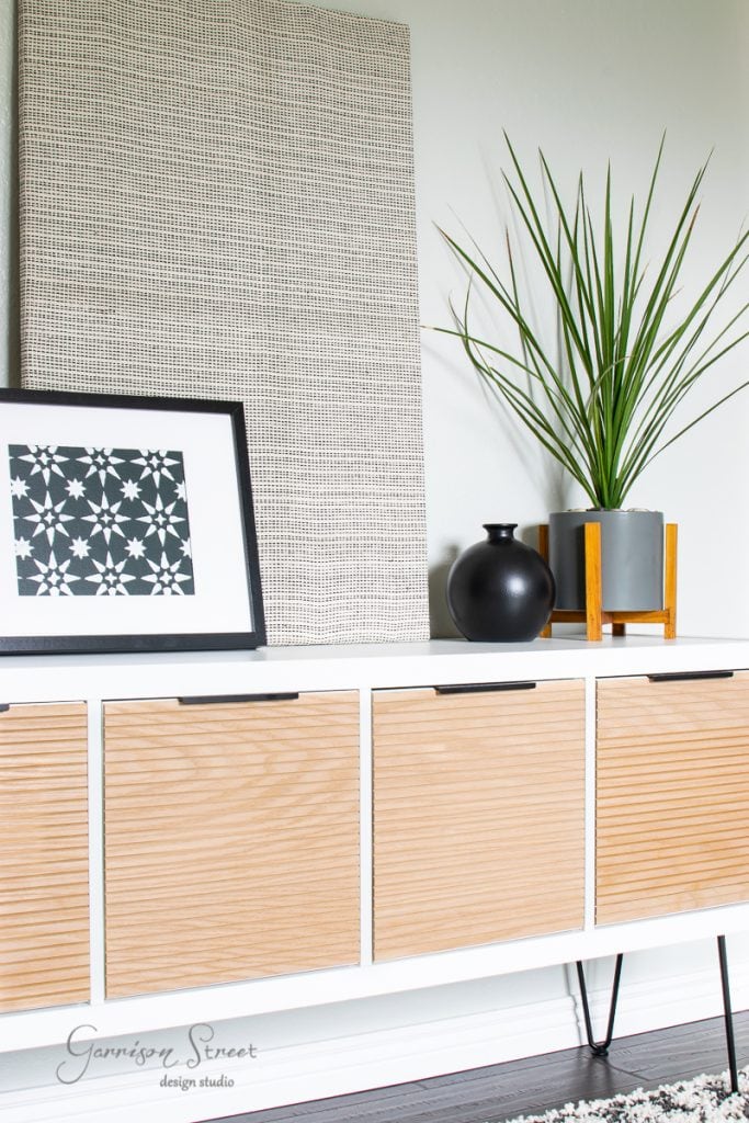 Home A modern sideboard perfect for a bunk room features light wood and white accents. On top, a black and white framed geometric print complements the small black vase and a potted plant with long, thin green leaves. A large textured panel leans stylishly against the wall behind them.