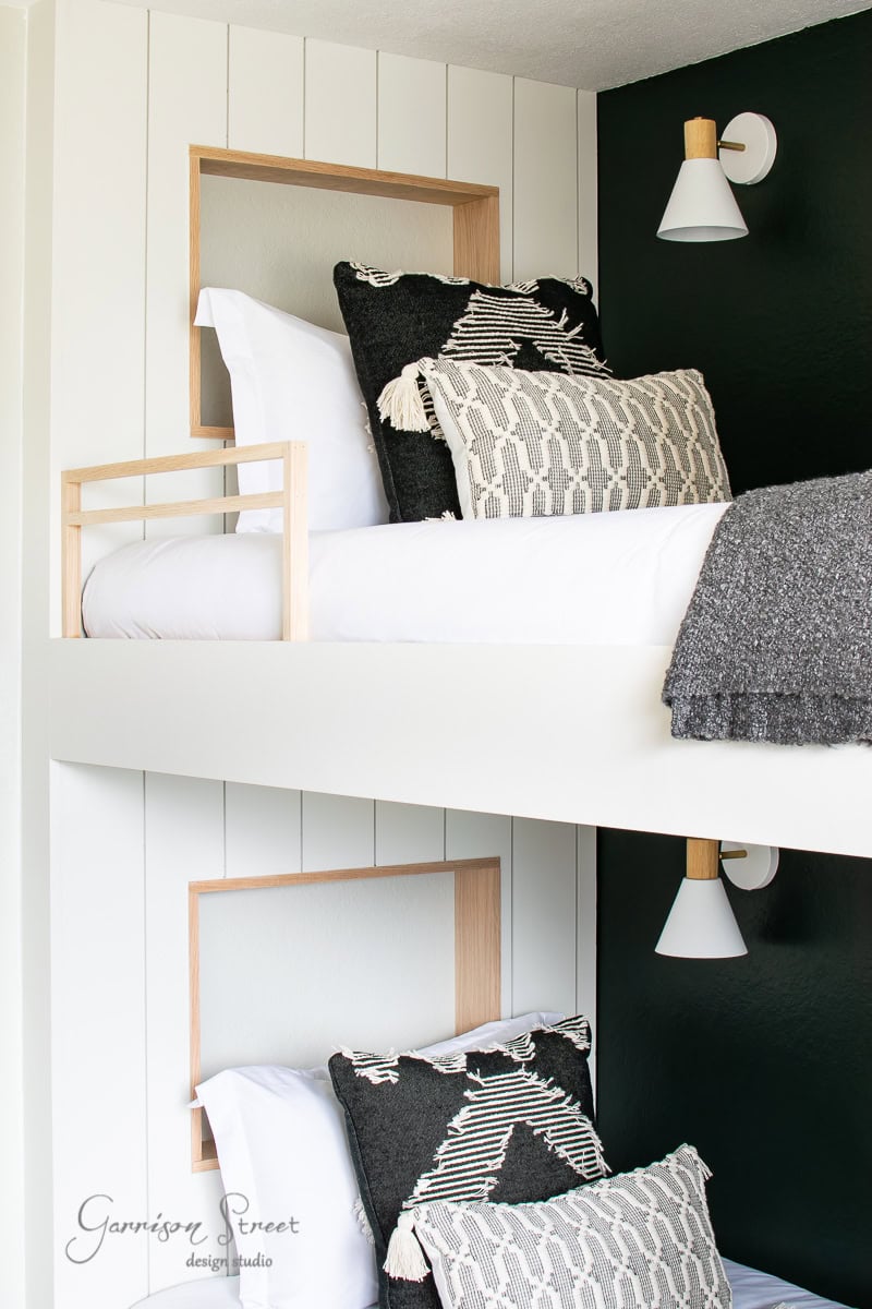 The bunk room boasts a modern bunk bed with wooden accents against white and dark walls. Each bed is adorned with black and white geometric-patterned pillows and dark gray blankets, while wall-mounted white lights are elegantly positioned above each bed.
