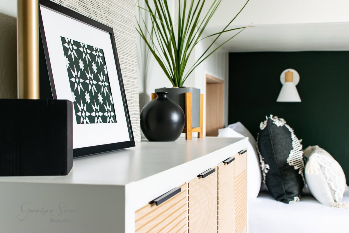 A modern bunk room features a sleek white dresser topped with a framed geometric print, a black vase, and decorative accents. The beds showcase black and white patterned pillows against a dark green accent wall with mounted white light fixtures.