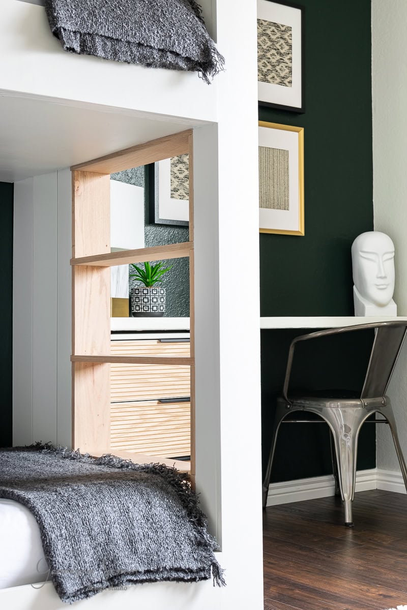 A modern bunk room with a bunk bed draped in gray blankets sits invitingly. A compact desk and chair rest against a dark green wall, beautifully adorned with framed art. Decorative touches include a sculpted head and a plant, complementing the sleek wooden floor.