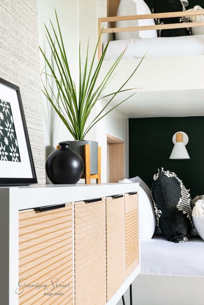 Modern interior featuring a wooden cabinet, green plant, and decorative black vase in a cozy bunk room. A bed with black and white cushions is visible above. A white wall light fixture adds to the minimalist design.