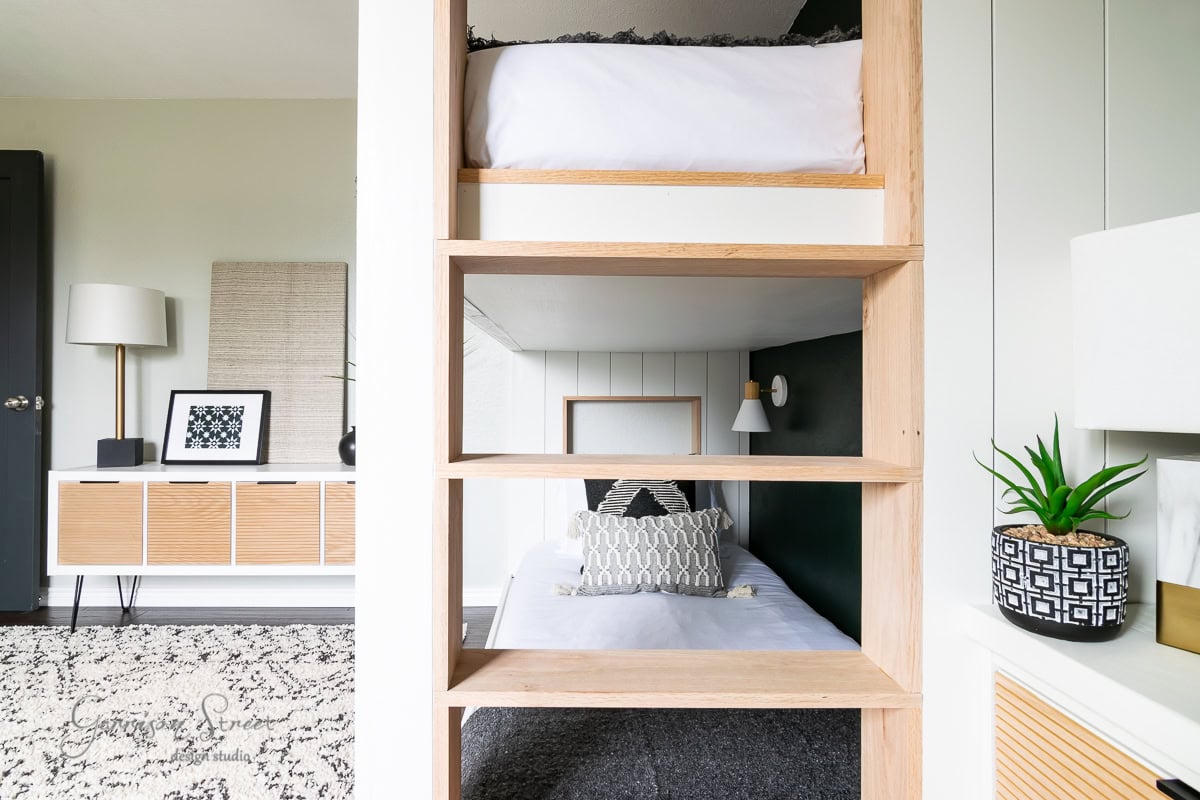 A modern bunk room features a bunk bed with wooden frames. The top bunk has white bedding, while the bottom is adorned with patterned pillows. A sideboard displaying decor items and a small potted plant rests against the light wall. A textured rug covers the floor.