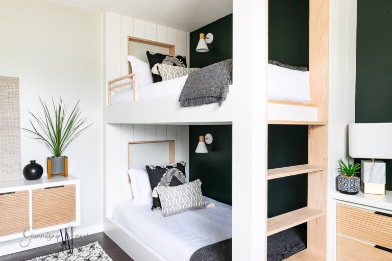 A sleek bunk room features a white and wood bunk bed against a dark green accent wall. It’s elegantly decorated with monochrome pillows and throws. To the left, a white dresser with wooden accents complements a potted plant, creating a stylish yet cozy atmosphere.