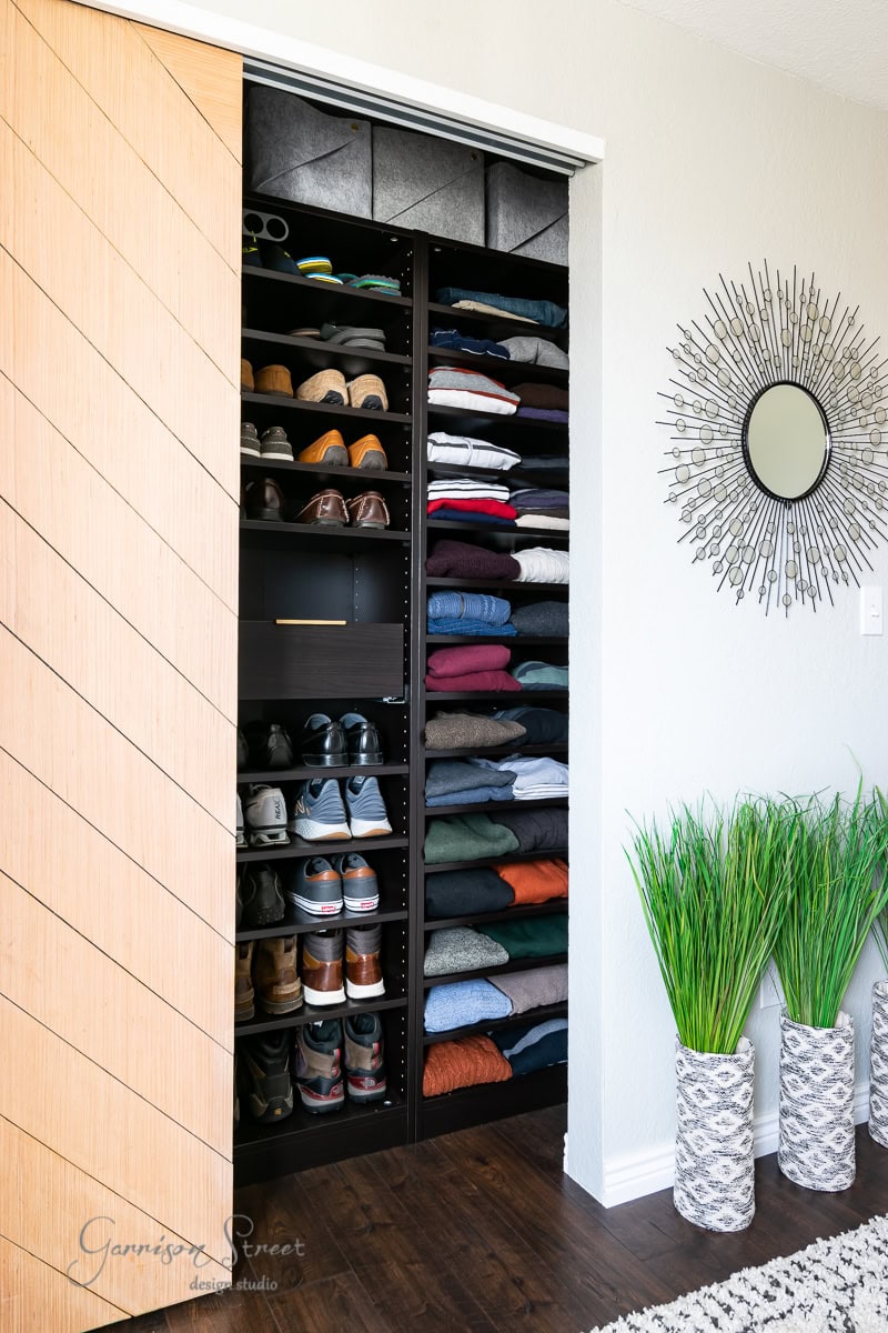 A neatly organized closet in the bunk room features shelves brimming with folded clothes, shoes, and hats. The closet boasts a wooden herringbone sliding door, while a sunburst mirror and potted plants adorn the adjacent wall and floor.