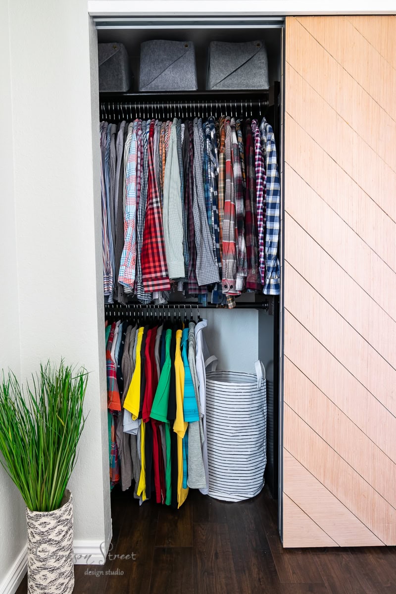 A closet in the bunk room with a wooden sliding door reveals neatly arranged clothing. Shirts hang on the upper rod, color-coordinated from light to dark. T-shirts are organized below. Gray storage bins occupy the top shelf, and a laundry basket is conveniently nearby.