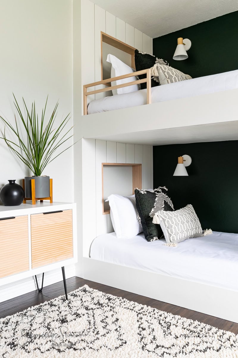 A sleek bunk room showcases two white bunk beds against a dark accent wall, each adorned with white and black patterned pillows. A mid-century style cabinet with potted plants rests on the dark wood floor, complemented by a decorative rug.