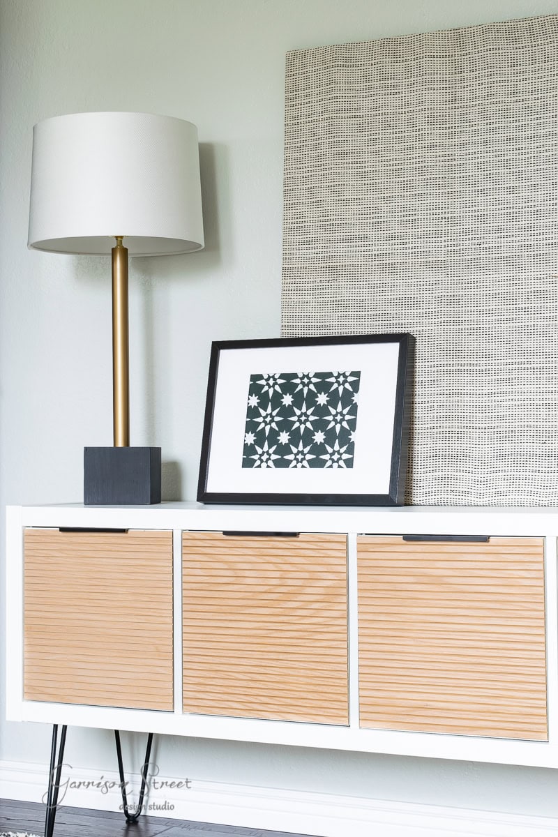 A sleek bunk room featuring a light wooden cabinet with three drawers, a tall lamp on a gold stand, and a framed geometric print. The light-colored walls complement the textured panel hanging behind the cabinet, creating an inviting and modern atmosphere.