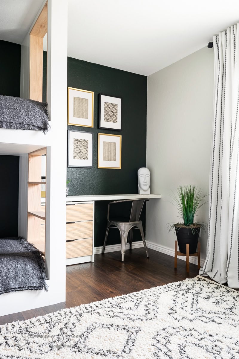 A modern bunk room features a sleek bunk bed and a compact desk area. The space showcases a dark accent wall adorned with framed art, while a white wall supports a floating desk with a chair and sculpted head. Nearby, a potted plant adds greenery, and a patterned rug adorns the wooden floor.