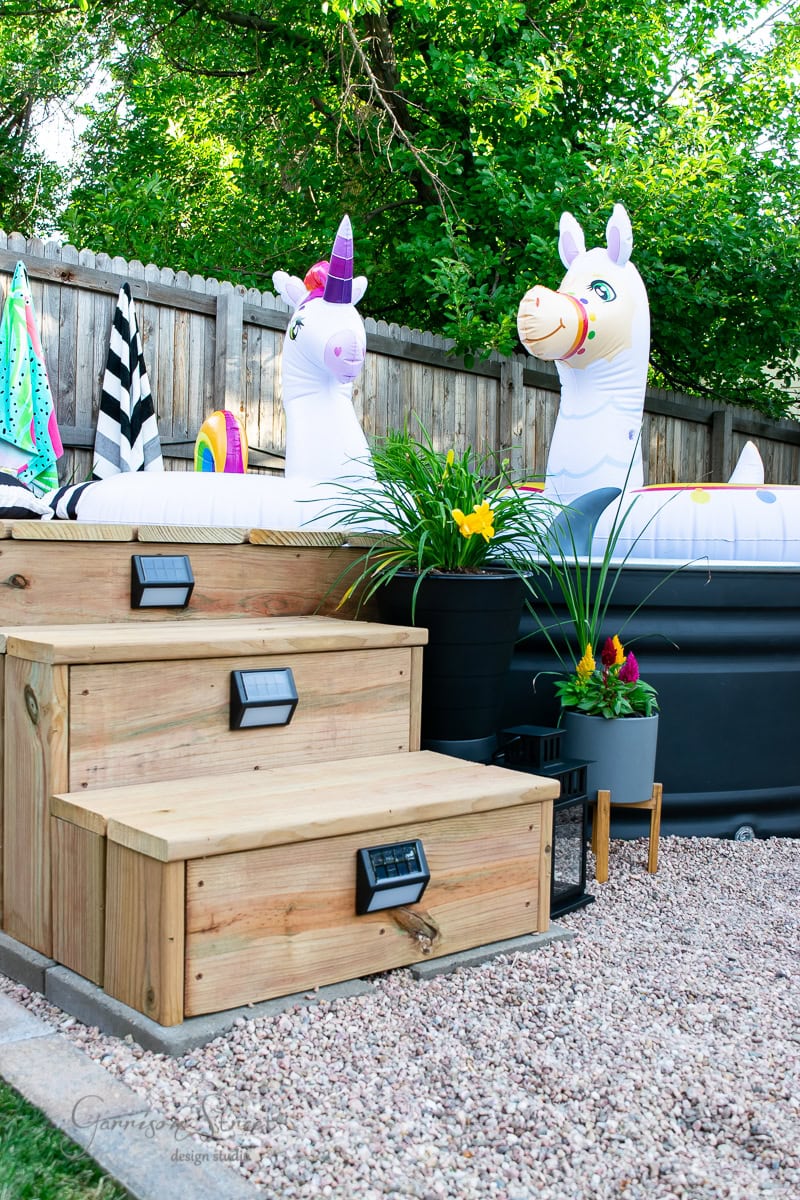 Wooden steps lead to an inflatable pool adorned with two unicorn floaties, earning it a spot in the "Best of 2021" backyard designs. A gravel path surrounds the area, bordered by a wooden fence and lush greenery. Potted plants with colorful flowers enhance the vibrant outdoor setting.