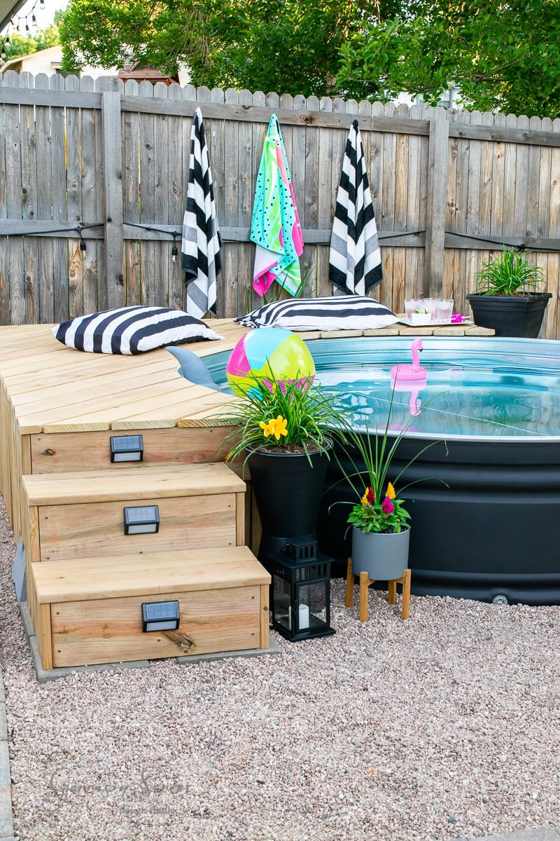 A small above-ground pool with clear water and flamingo floats is surrounded by a wooden deck, reminiscent of 2021 summer vibes. Striped towels hang on a fence, while potted plants and balloons add charm to this "Best of" backyard retreat. Wooden steps gracefully lead up to the inviting deck.