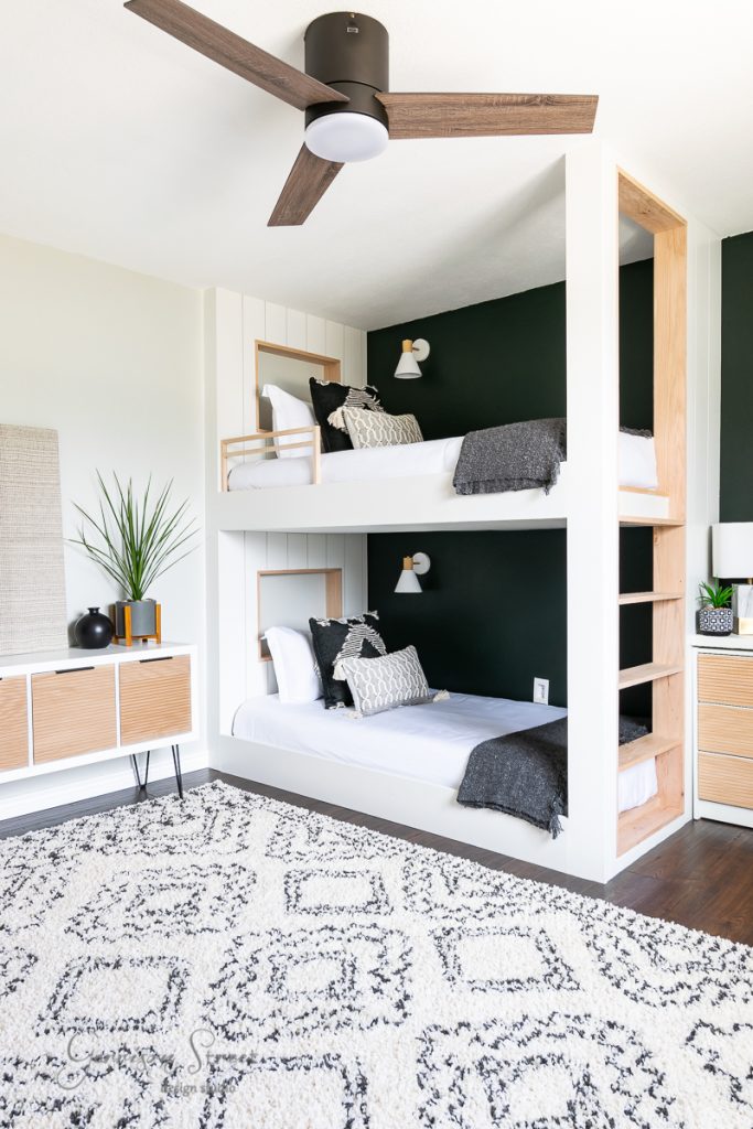 A modern, cozy bedroom showcasing the best of 2021 design with two white bunk beds against a dark green accent wall. Each bed is adorned with black and white bedding. The room includes sleek wooden furniture, a patterned rug, and a ceiling fan with elegant wooden blades.