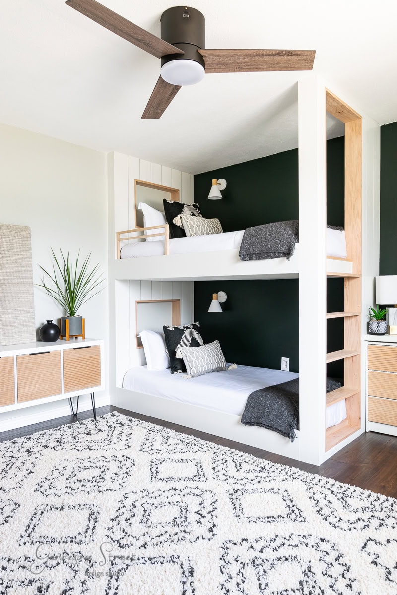 A modern, cozy bedroom showcasing the best of 2021 design with two white bunk beds against a dark green accent wall. Each bed is adorned with black and white bedding. The room includes sleek wooden furniture, a patterned rug, and a ceiling fan with elegant wooden blades.