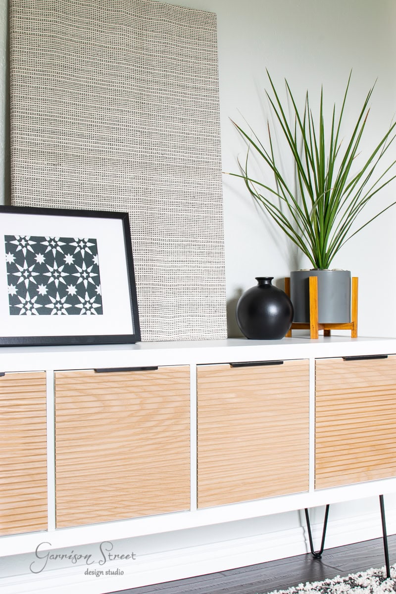A modern sideboard with wooden doors and black metal legs, part of the Best of 2021 collection. On top, there's a decorative plant in a gray pot with a wooden stand, a black vase, and framed artwork featuring a geometric star pattern. A textured wall panel serves as the backdrop.