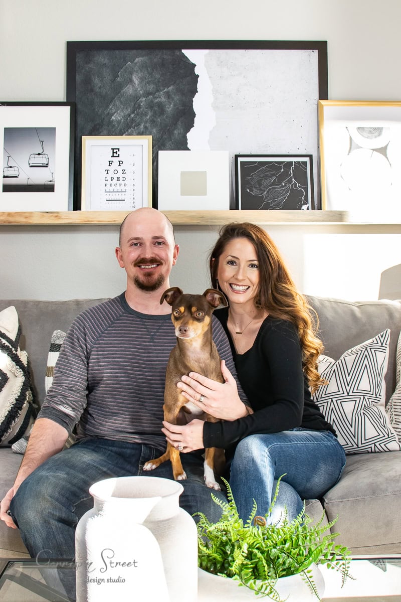A smiling couple sits on a couch in a stylish living room, holding the best of 2021—a small brown dog. The room features modern art on the walls, patterned throw pillows, and a vase with greenery on the coffee table.